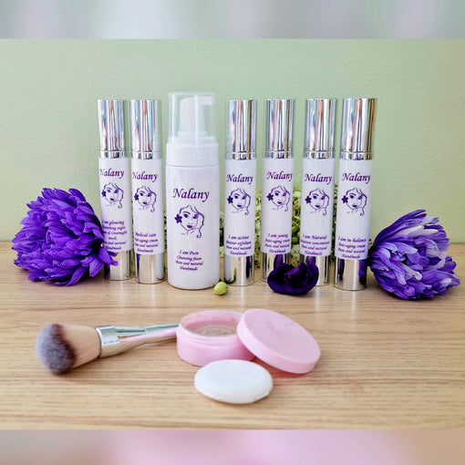 Foto met de producten van Nalany skincare in witte flacons met zilveren dopjes en paars logo vergezeld met twee prachtige paarse bloemen en op de voorgrond de minerale foundation met brush 